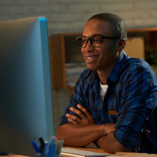 Handsome young man sitting in front of computer with arms crossed and watching his favorite movie, interior of dark bedroom on background
