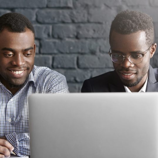 Two happy African colleagues using laptop. Manager in shirt showing presentation on notebook computer to his boss wearing suit and glasses, during meeting in modern office interior with brick walls